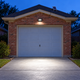Small 300-LED solar floodlight mounted above a brick garage door, with a separate rooftop solar panel illuminating the driveway at dusk.