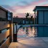 A professional lifestyle photograph of a modern black aluminum LED bollard light (60cm) mounted on a rustic wood fence beside a residential swimming pool during twilight, featuring realistic light reflections on water, textured concrete pavers, and pool house for scale. Realistic style, shot on Nikon Z5II.
