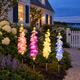 Four pastel solar delphinium stake lights beside a shell gravel path and white picket gate in a Cape Cod coastal cottage garden at blue hour.