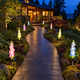Eight solar delphinium flower lights spaced along both sides of a winding garden walkway beside a Pacific Northwest cedar house at night, with clear walking space, exposed silver stems and upward-facing solar panels.