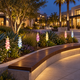 Solar delphinium flower lights accent a curved planting bed behind wooden public seating in a San Diego pedestrian plaza at blue hour.